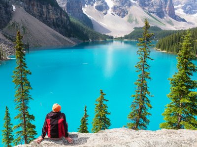 Bright turquoise blue Moraine lake being admired by person sat down on rock with tall green pine trees and mountains surrounding lake at Banff National Park, Canada