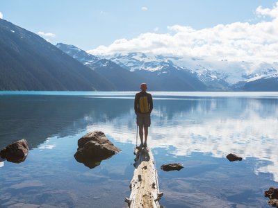 Hiker standing on floating log on edge of Garibaldi Lake admiring view of snow-capped mountains with reflection from lake near Whistler, British Columbia, Canada